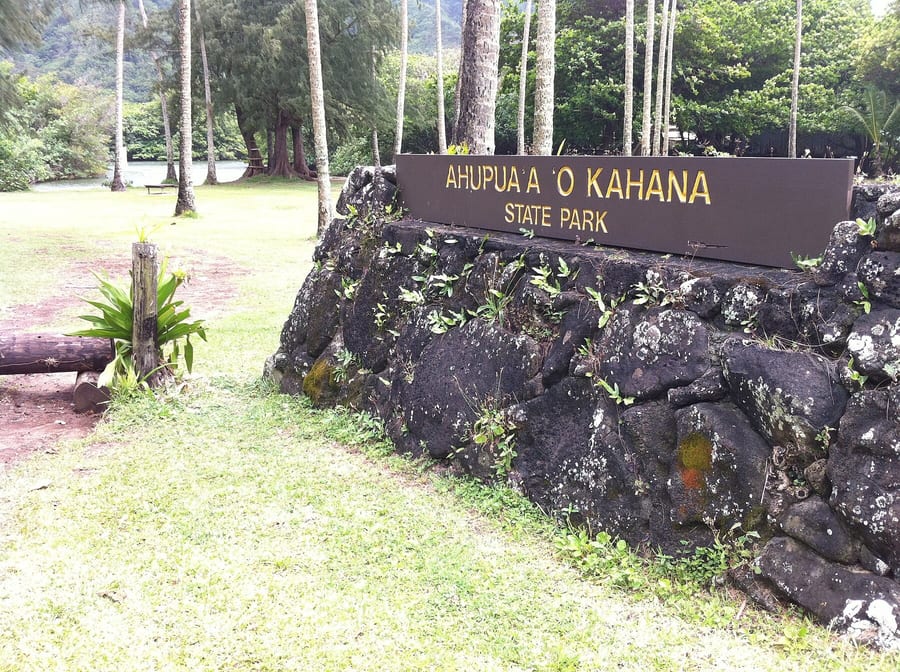 Wooden sign reading Ahupuaʻa O Kahana State Park on a lava-rock base, with lawn and trees behind it.